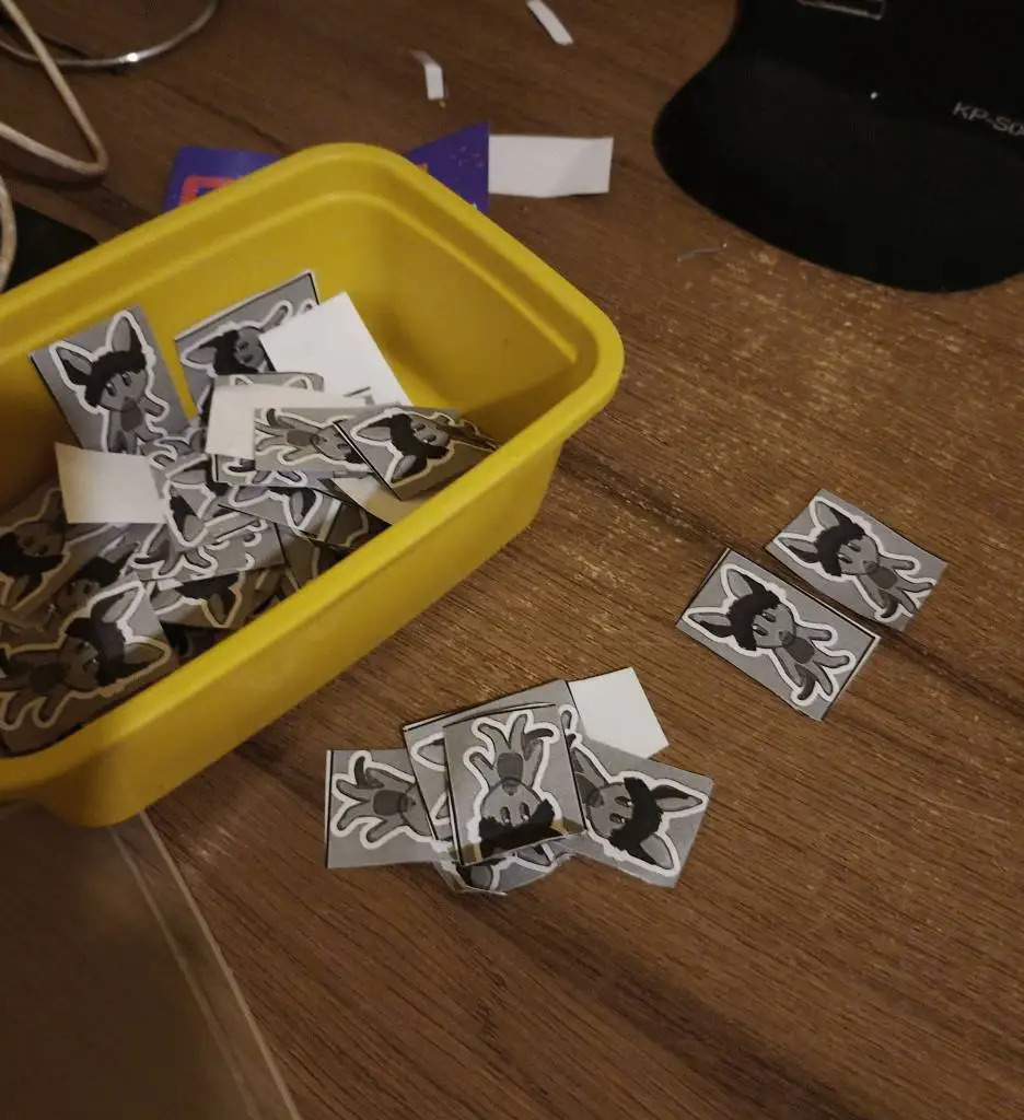 A large heap of grayscale Gus the Wallaby stickers in rectangular cutouts, some on a wooden desk, most inside a small yellow plastic container.