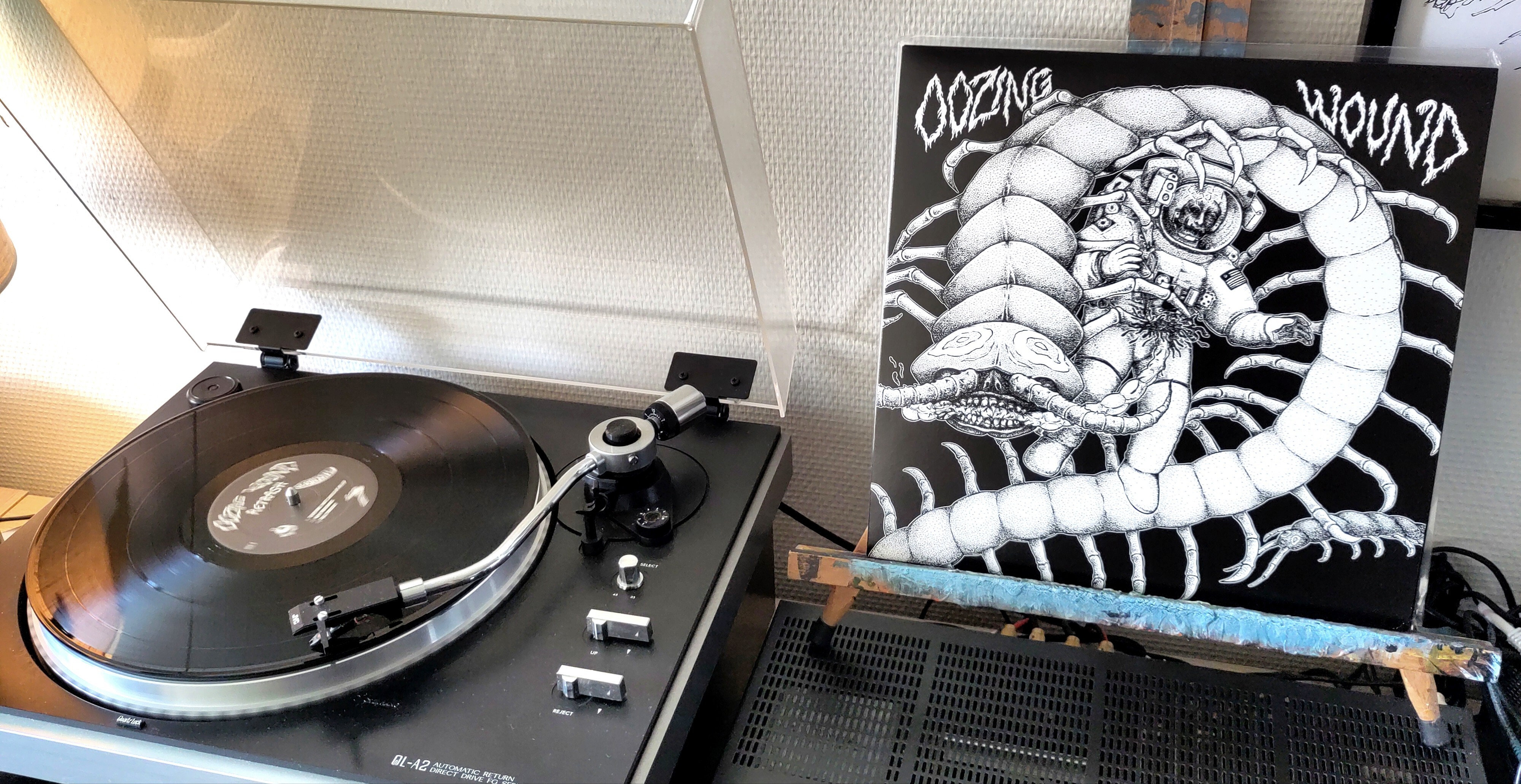 Left: the black vinyl spinning on the turntable. Right: the LP cover stands on an easel. The cover depicts an astronaut being gored by a giant centipede. It's rendered in black and white pen drawing.